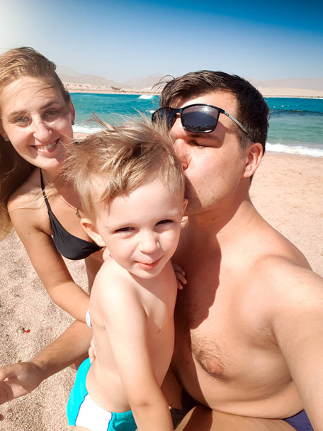 Selfie portret rodziców całuje małego malucha syna na plaży morskiej w słoneczny wietrzny dzień. Rodzinny relaks i dobry czas podczas letnich wakacji. - Zdjęcie, obraz
