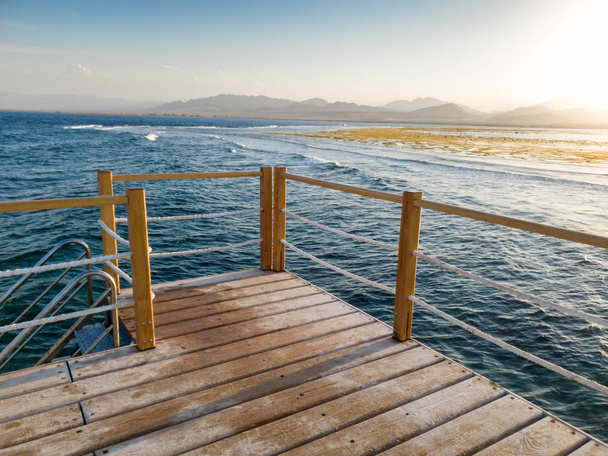 Prachtig landschap van lange houten pier en kalme golven van de zee tegen zonsondergang en bergen - Foto, afbeelding