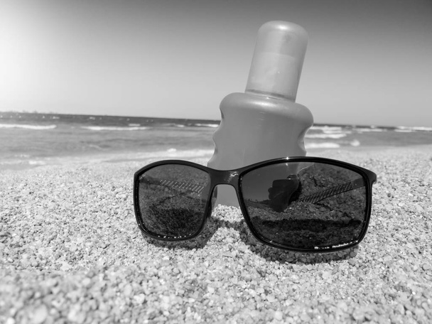 Czarno-biały wizerunek okularów przeciwsłonecznych i kremu ochronnego leżącego na piaszczystej plaży na morzu. Doskonały obraz do ilustrujących letnie wakacje. - Zdjęcie, obraz