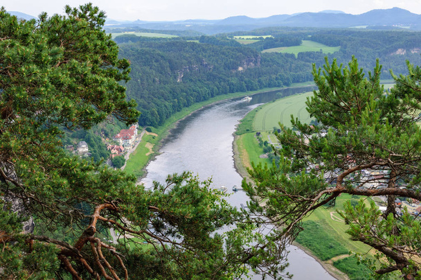 Aerial view of the navigable river among the green forests.Picturesque tree branches in the foreground, the ship sails on the river.Houses with red roofs on the shore. - Фото, изображение