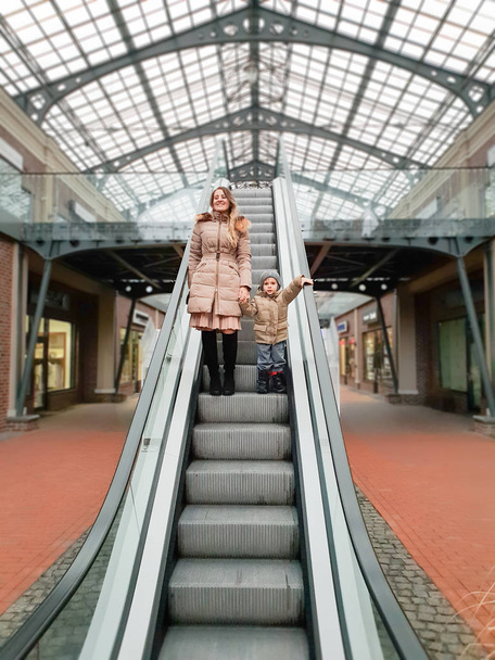 schöne lächelnde junge Mutter hält ihren kleinen Sohn an der Hand, während sie auf der Rolltreppe im Einkaufszentrum steht - Foto, Bild