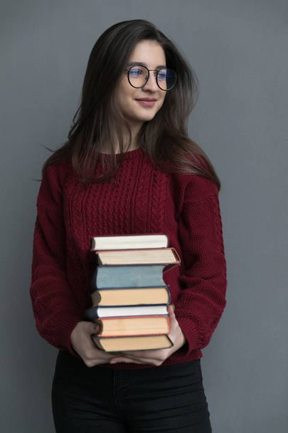 Primo piano di una ragazza con i capelli scorrevoli, su uno sfondo grigio che tiene i libri nelle sue mani, posa. Indossare un maglione bordeaux e occhiali. - Foto, immagini