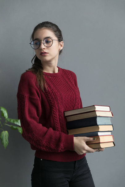 Closeup of a girl with collected hair, on a gray background holding books in her hands. Wearing a burgundy sweater and glasses. - Photo, image