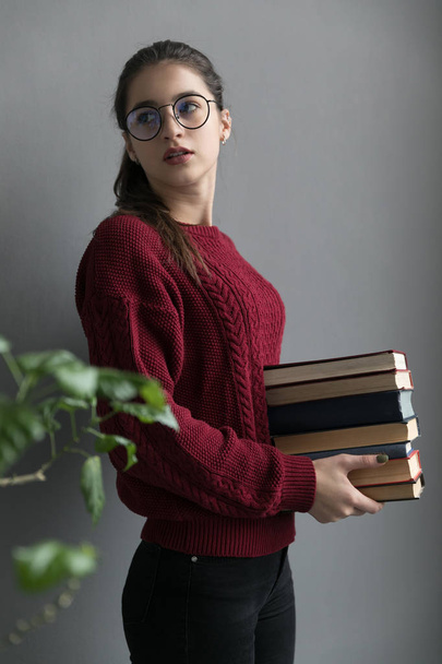 Closeup of a girl with collected hair, on a gray background holding books in her hands. Wearing a burgundy sweater and glasses. - Fotografie, Obrázek