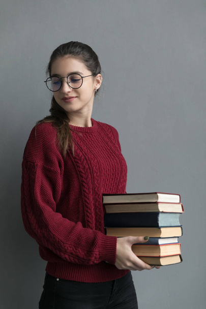 Detailní záběr dívky se sebranými vlasy, na šedém pozadí, držící knihy v rukou. Na sobě má burgundský svetr a brýle. - Fotografie, Obrázek