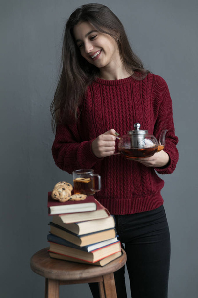 A close-up of the book stacked one on top of the other, on the books are cookies and a cup of tea, the girl is wearing a red sweater, she is holding a tea bar with a gray background. - Foto, Imagen