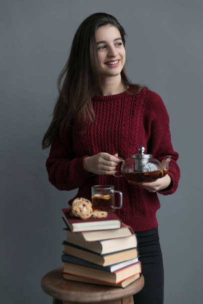 A close-up of the book stacked one on top of the other, on the books are cookies and a cup of tea, the girl is wearing a red sweater, she is holding a tea bar with a gray background. - Fotó, kép