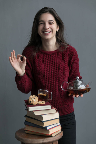 A close-up of the book stacked one on top of the other, on the books are cookies and a cup of tea, the girl is wearing a red sweater, she is holding a tea bar with a gray background. - Fotoğraf, Görsel