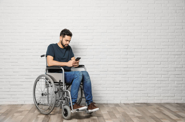Young man with smartphone in wheelchair near brick wall indoors. Space for text - Фото, изображение