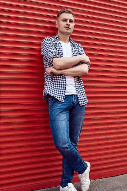 Jovem homem bonito em uma camisa e jeans perto do portão vermelho
 - Foto, Imagem