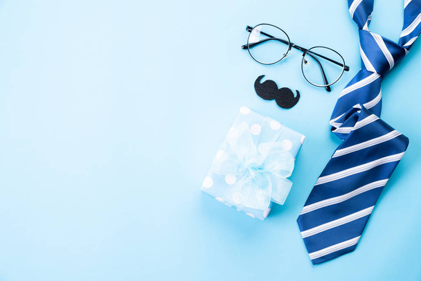 Happy fathers day concept. Top view of blue tie, beautiful gift box, glasses and mustache on bright blue pastel background. Flat lay. - Photo, image