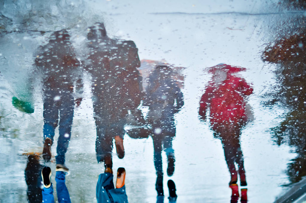 Reflexão turva em uma poça de quatro pessoas andando na rua molhada da cidade durante a chuva e neve. Conceito de humor
 - Foto, Imagem