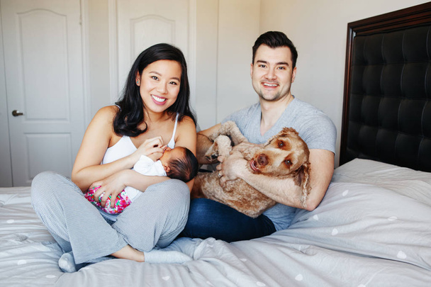 Усміхнені китайські азіатські мати і кавказькі батько з змішаною раси новонароджених немовляти сина дочки і великі собаки Pet. Щаслива родина в спальні. Головна стиль життя автентичний природний момент.  - Фото, зображення