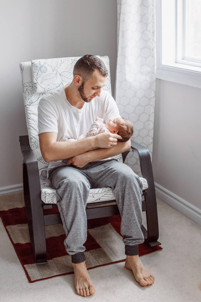 Caucasian father sitting in armchair with newborn baby girl. Parent holding rocking child daughter on his hands. Authentic lifestyle documentary moment. Single dad family life concept. - Photo, Image