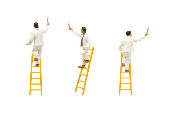 Miniature figurine character as painter standing on wooden ladder and painting wall with paint tools isolated on white background. - Photo, Image