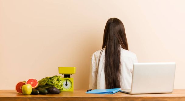 Young nutritionist chinese woman working with her laptop from behind, looking back. - Фото, зображення