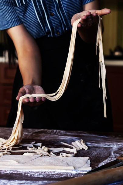 Молодая домохозяйка готовит домашнюю пасту на кухне. Женские руки держат длинную полоску теста для приготовления макарон
. - Фото, изображение
