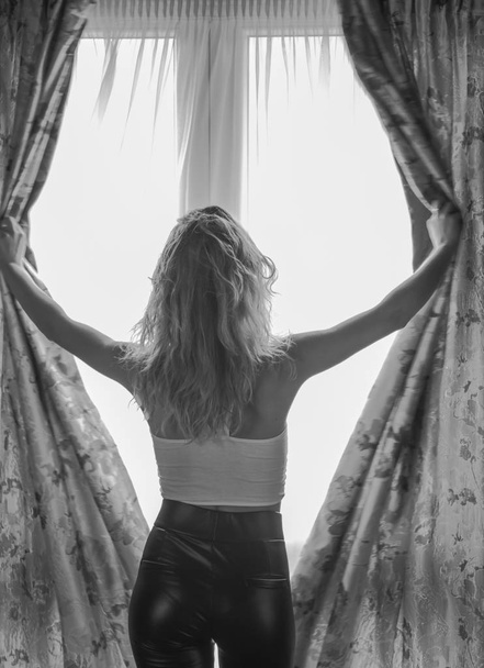 Back view of blonde young woman opening curtains on wide window - Foto, imagen