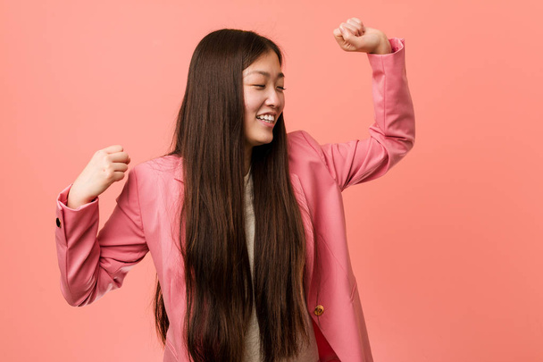 Joven mujer china de negocios vistiendo traje rosa celebrando un día especial, salta y levanta los brazos con energía
. - Foto, Imagen