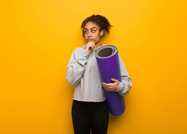 Young fitness black woman doubting and confused. Holding a mat. - 写真・画像