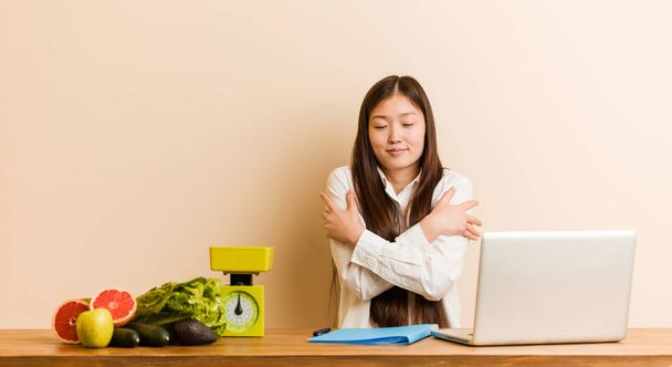 Jovem nutricionista mulher chinesa trabalhando com seu laptop abraça a si mesmo, sorrindo despreocupado e feliz
. - Foto, Imagem