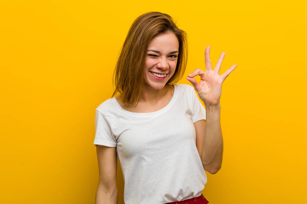 Young natural caucasian woman winks an eye and holds an okay gesture with hand. - Photo, Image