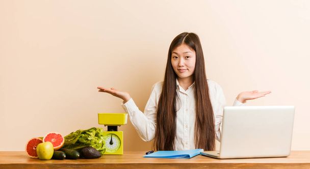 Jovem nutricionista mulher chinesa trabalhando com seu laptop duvidando e encolhendo os ombros no gesto de questionamento
. - Foto, Imagem
