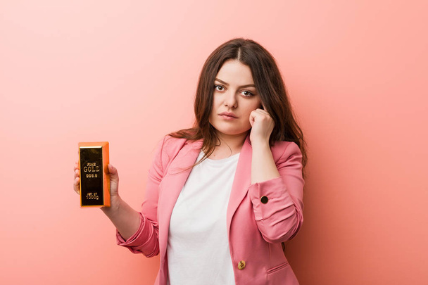 Young plus size curvy business woman holding a gold ingot pointing his temple with finger, thinking, focused on a task. - Photo, Image