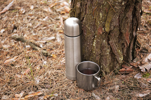 Thermos and mug of tourist near tree in forest - Photo, Image
