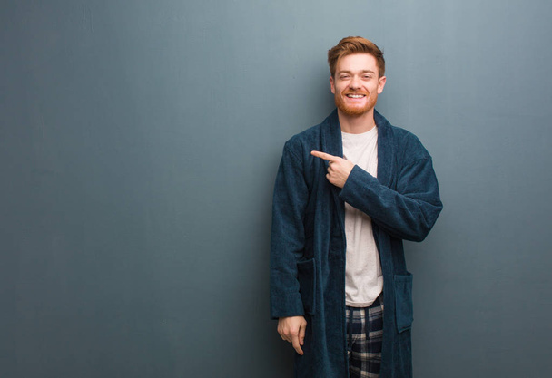 Young redhead man in pajama smiling and pointing to the side - 写真・画像