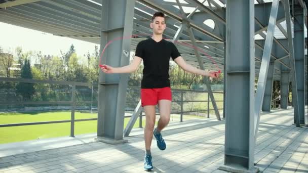 Portrait Of Fit Young Man With Jump Rope On Platform Near Metal Racks. Fitness Skipping Workout Outdoors. The Guy Jumps Near The Metal Pillars In The Background Of The Stadium. Dressed In A Black T - Filmmaterial, Video