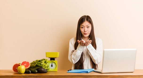 Jovem nutricionista mulher chinesa trabalhando com seu laptop lábios dobráveis e segurando palmas para enviar beijo de ar
. - Foto, Imagem