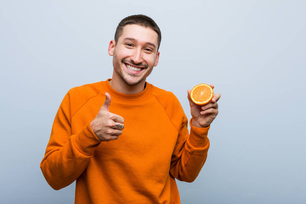 Jovem caucasiano segurando uma laranja sorrindo e levantando o polegar
 - Foto, Imagem