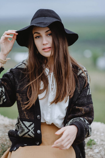 Young woman in knitted coat and black hat posing sitting on top of hill - Photo, Image