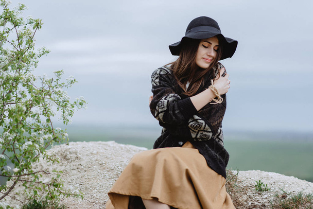 Fiatal nő kötött kabátban és fekete kalapban pózol a domb tetején ülve ölelkező vállakkal - Fotó, kép