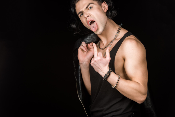 young man showing middle finger and tongue while holding leather jacket isolated on black  - Fotografie, Obrázek