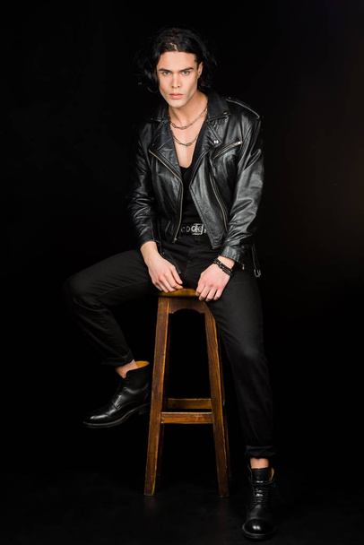 серйозна людина в шкіряній куртці, дивлячись на камеру, сидячи на стільці ізольовано на чорному
  - Фото, зображення