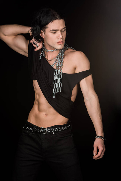 handsome man with chains around neck touching tank top while standing isolated on black  - Zdjęcie, obraz