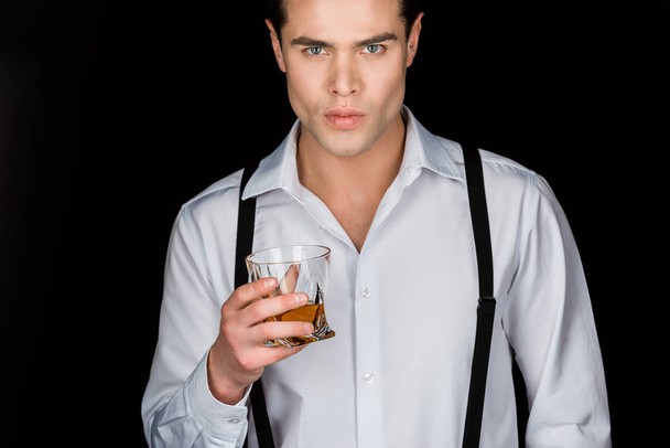 handsome man in white shirt and suspenders holding glass of whiskey isolated on black  - Valokuva, kuva