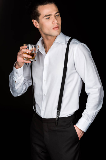 handsome man in white shirt and suspenders holding glass of whiskey while standing with hand in pocket isolated on black  - Foto, Imagen
