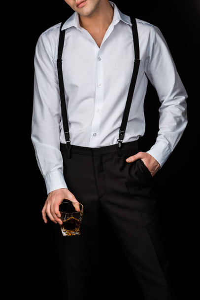 vista cortada do homem em camisa branca e suspensórios segurando vidro de uísque enquanto em pé com a mão no bolso isolado em preto
  - Foto, Imagem