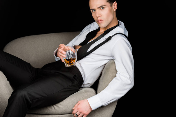serious man in white shirt and suspenders holding glass of whiskey while sitting in armchair isolated on black  - Foto, immagini