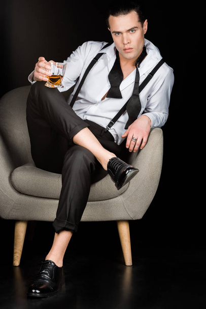σοβαρός άνθρωπος κρατώντας ένα ποτήρι ουίσκι και καθισμένος στην πολυθρόνα σε μαύρο  - Φωτογραφία, εικόνα
