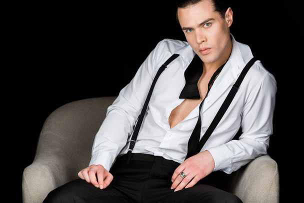 confident man in white shirt and suspenders sitting in armchair isolated on black  - Foto, Imagen