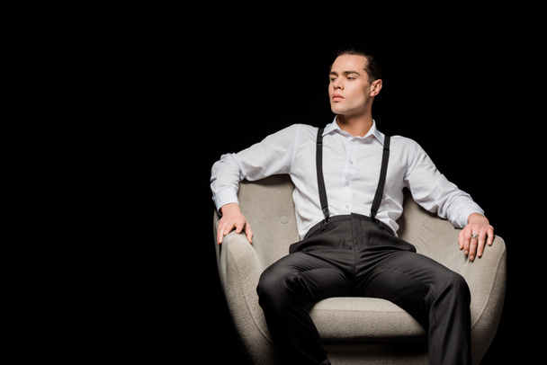 handsome man in shirt sitting in armchair isolated on black  - Fotó, kép