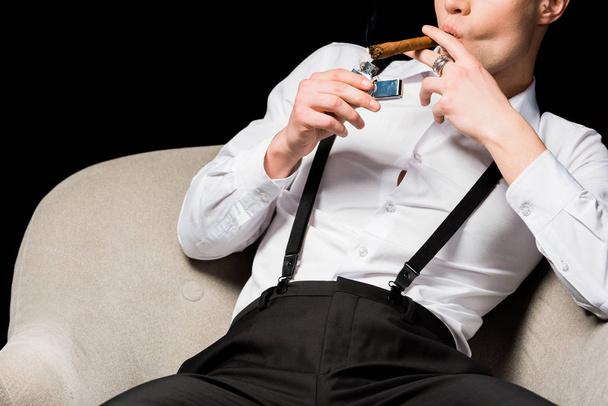 cropped view of man holding lighter near cigar while sitting in armchair isolated on black  - Φωτογραφία, εικόνα
