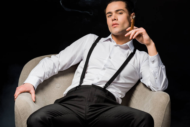 homme sérieux tenant cigare tandis que assis dans un fauteuil isolé sur noir
  - Photo, image