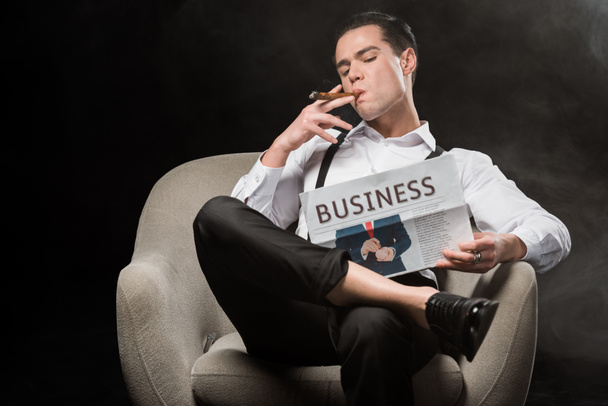 selective focus of handsome man sitting in armchair reading business newspaper and smoking cigar isolated on black - Foto, Imagen