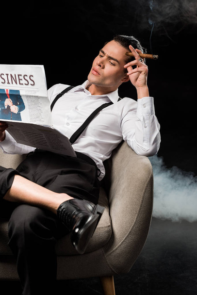 selective focus of handsome man sitting in armchair reading business newspaper and holding cigar on black with smoke  - Foto, Imagen