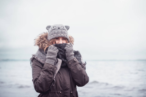 Pretty squinting girl in warm winter clothing spending time at the beach. - Photo, image